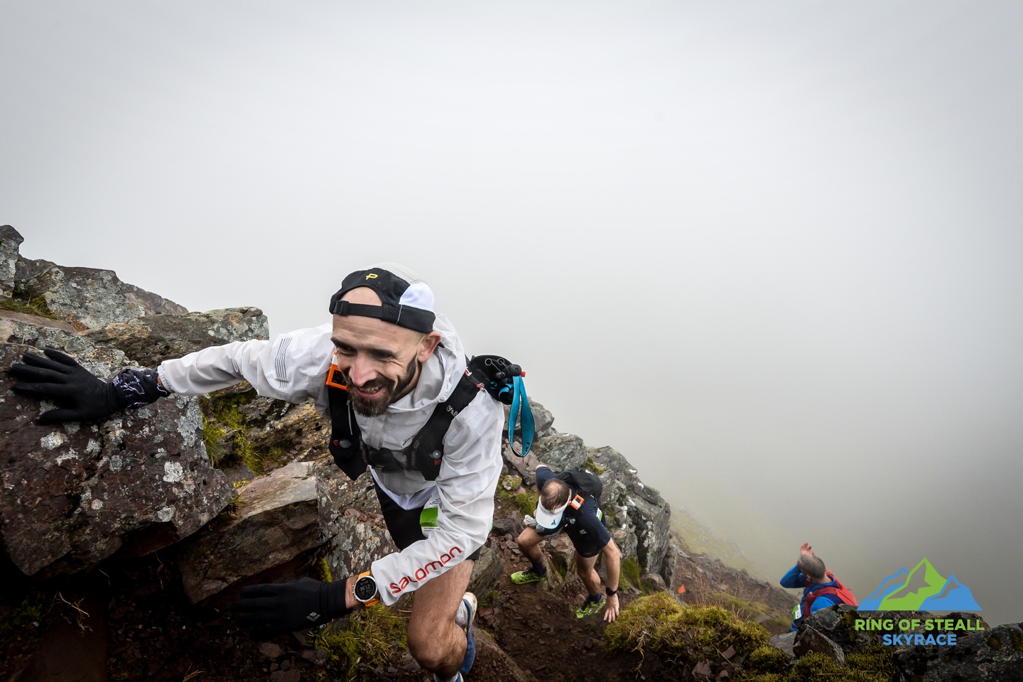 Pete grabs a rock for support while scrambling up a steep section of mountain side. Two other runners can be seen below him, with mist obscuring the view further away.