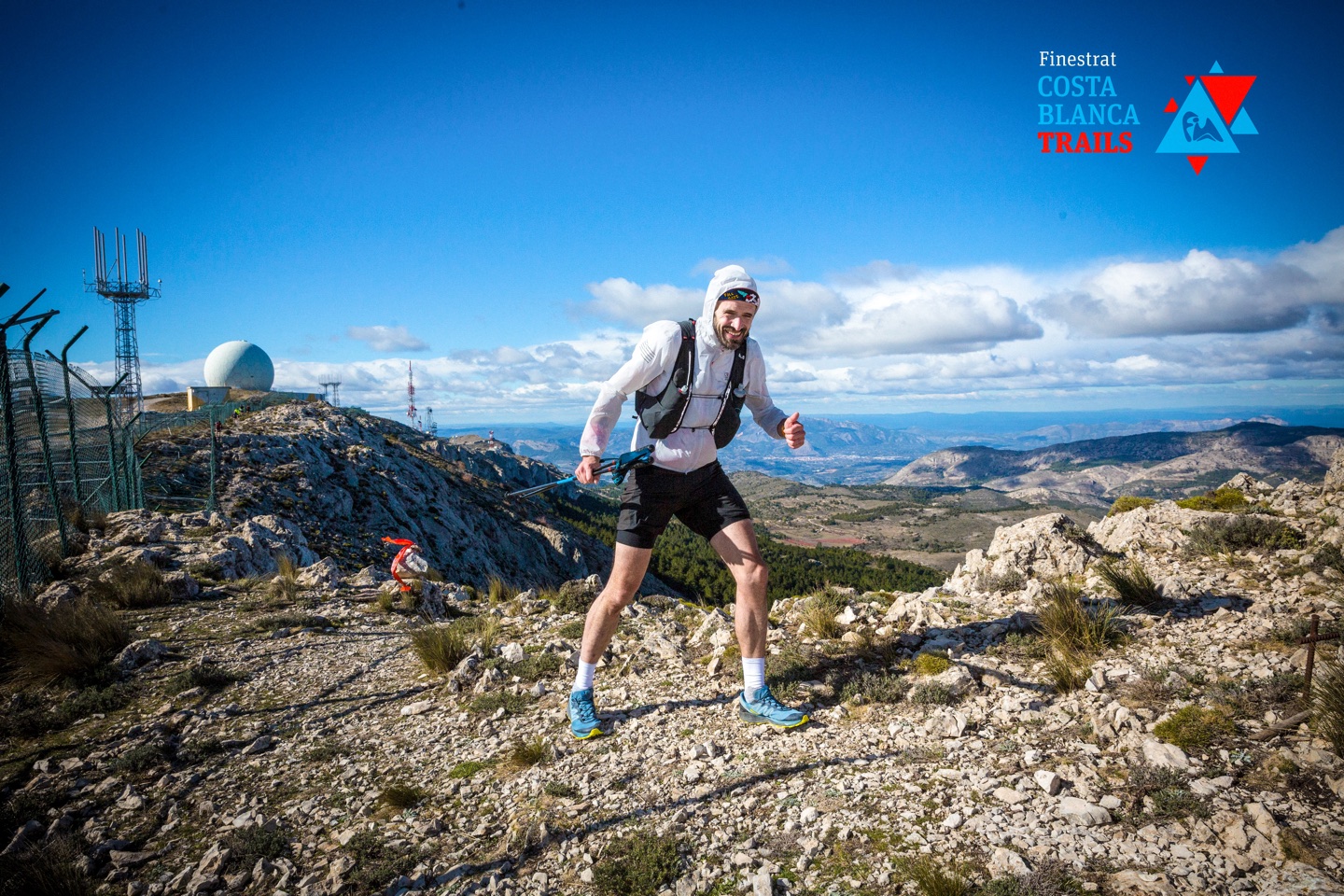 A runner in a white waterproof with the hood up, and a black hydration pack, braces against the wind at the top of a mountain. The sky is blue with few clouds. A radar dome is visible in the background and the security fence of a air force base runs along the left of the picture.