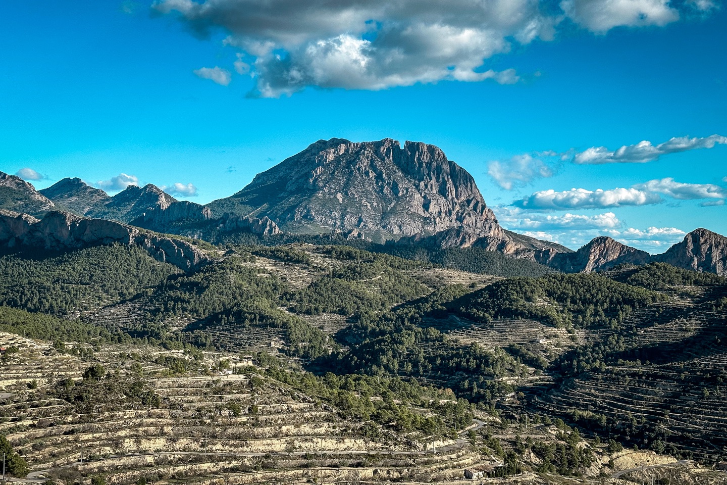 The mountain of Puig Campana is seen from the north, with rolling foothills of greeney in front of it. The sky is a bright blue colour with a handful of clouds above the peak itself.