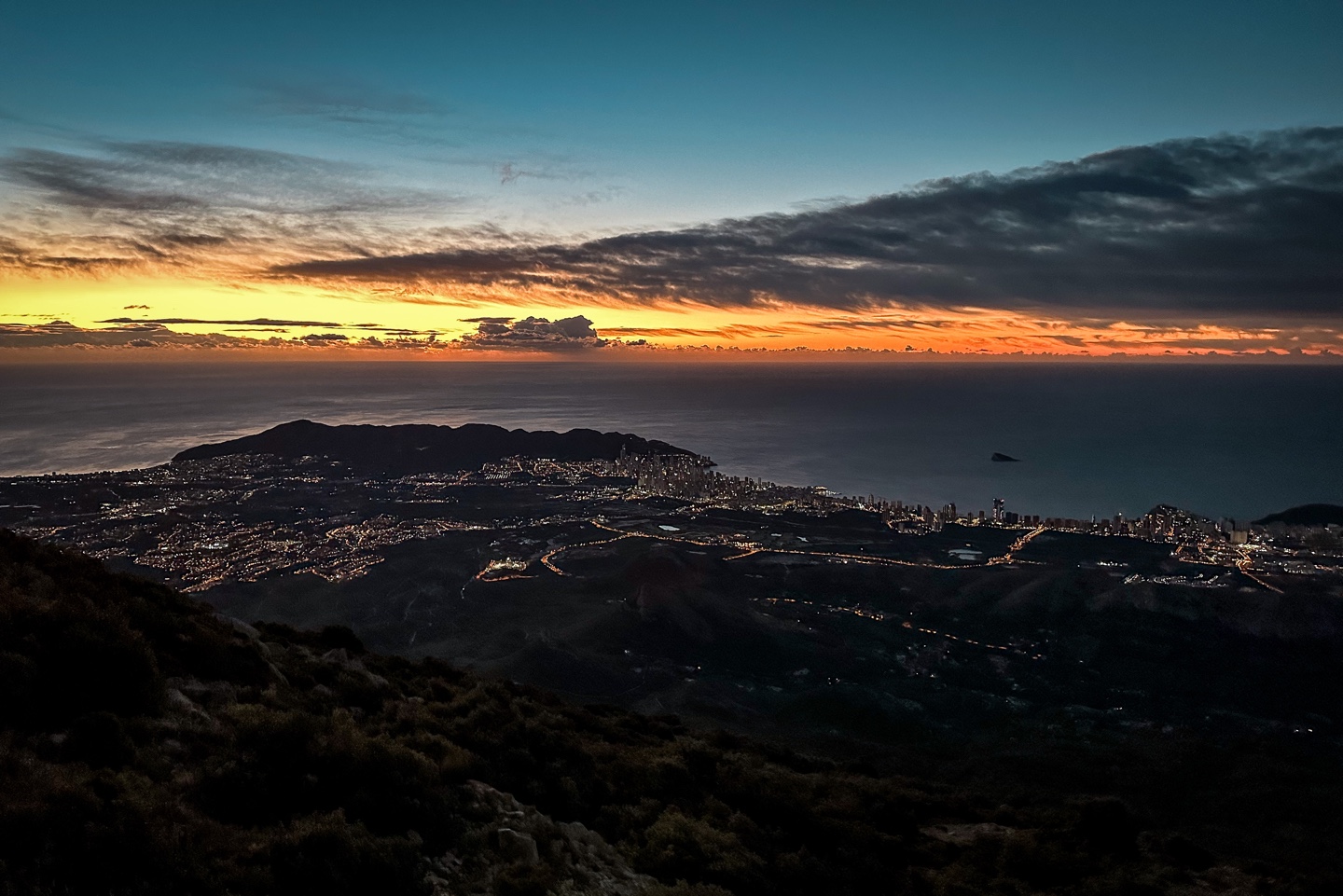 The view from the summit of a mountain (Puig Campana) at sunrise. The Mediterranean Sea is in the background with the first orange light appearing on the horizon. In the foreground the land appears black with only the lights of the coastal town below visible.