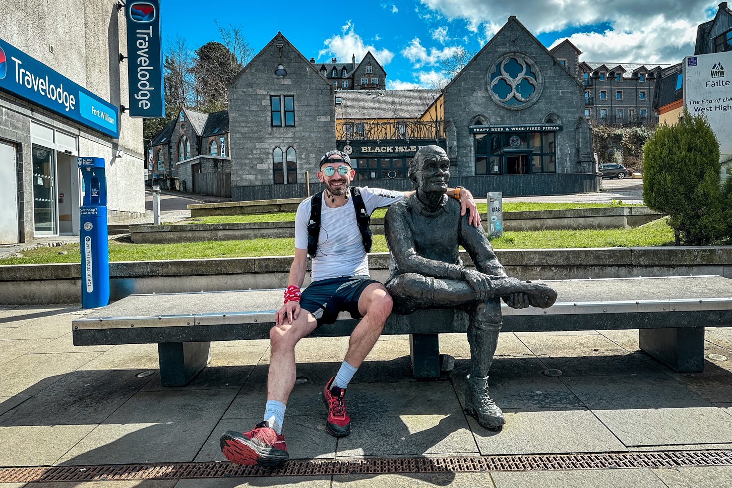 Pete sat next to the West Highland Way hiker's statue in Fort William. It's a sunny day and Pete is smiling wearing red running shoes, black shorts and a white top.