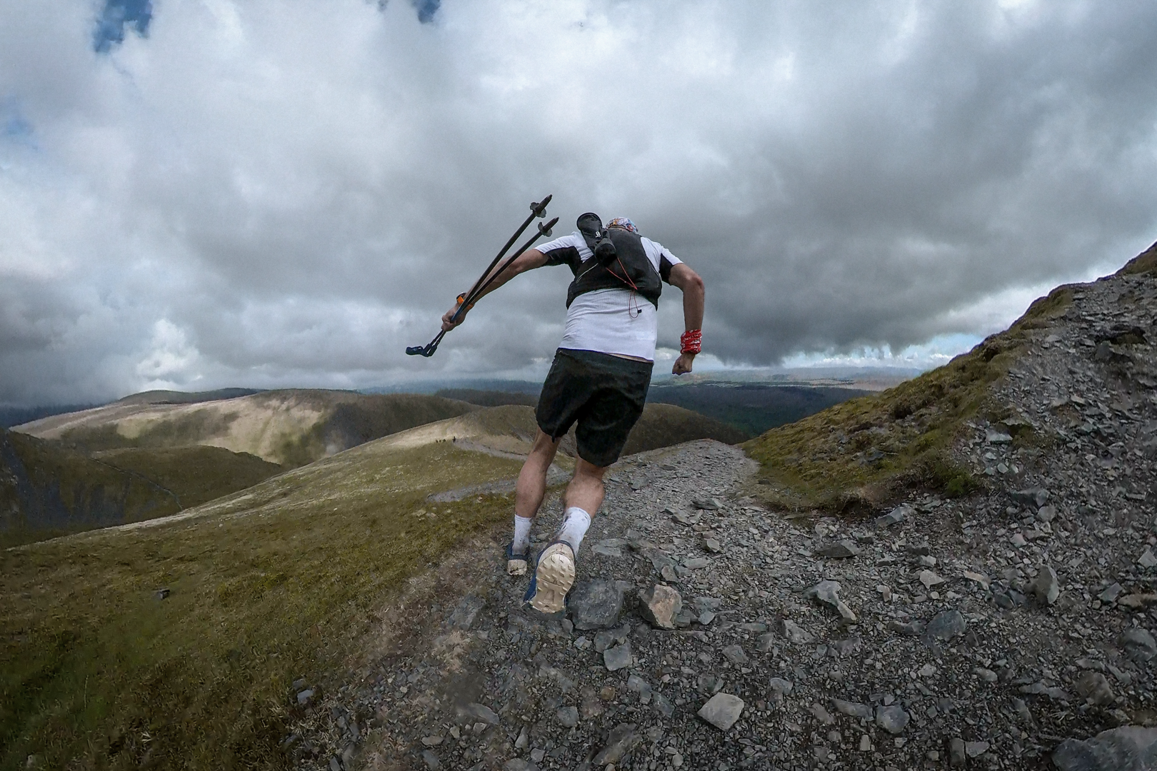 Pete is running down the swithcback of a rocky path on the side of a mountain. His feet are off the ground and he's leaning forward, gripping trekking poles in his left hand. He's wearing a white top and black shorts.