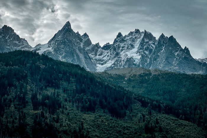A series of sharp mountain summits, with snow on the north faces, and a green pine forest in the foothills.