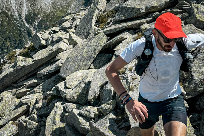 A man in a white tee-shirt and orange hat runs towards the camera over a boulder field.