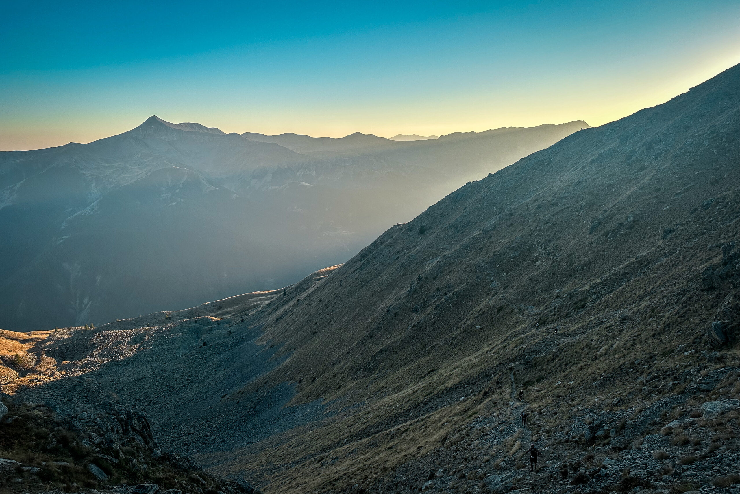 The evening light filters through a valley, emerging from the shoulder of one mountain and casting it's glow across the mountains beyond. In the foreground, almost lost in the scale, are two runners.
