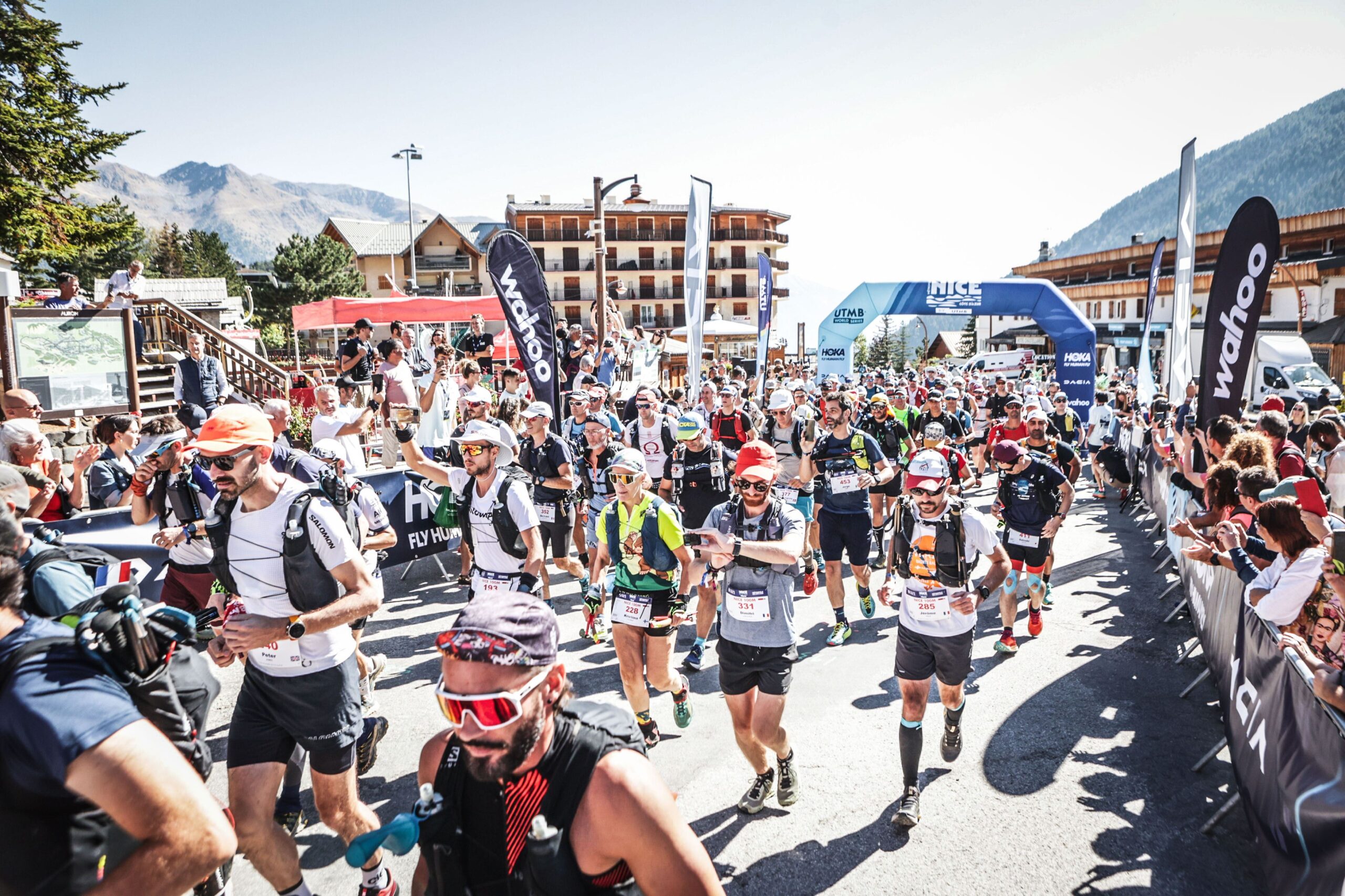 A number of trail runners, wearing hydration vests, are running across the startline of a race. They have just run through an arch that says 'Nice by UTMB', the sun is shining and the setting is an alpine town in France.