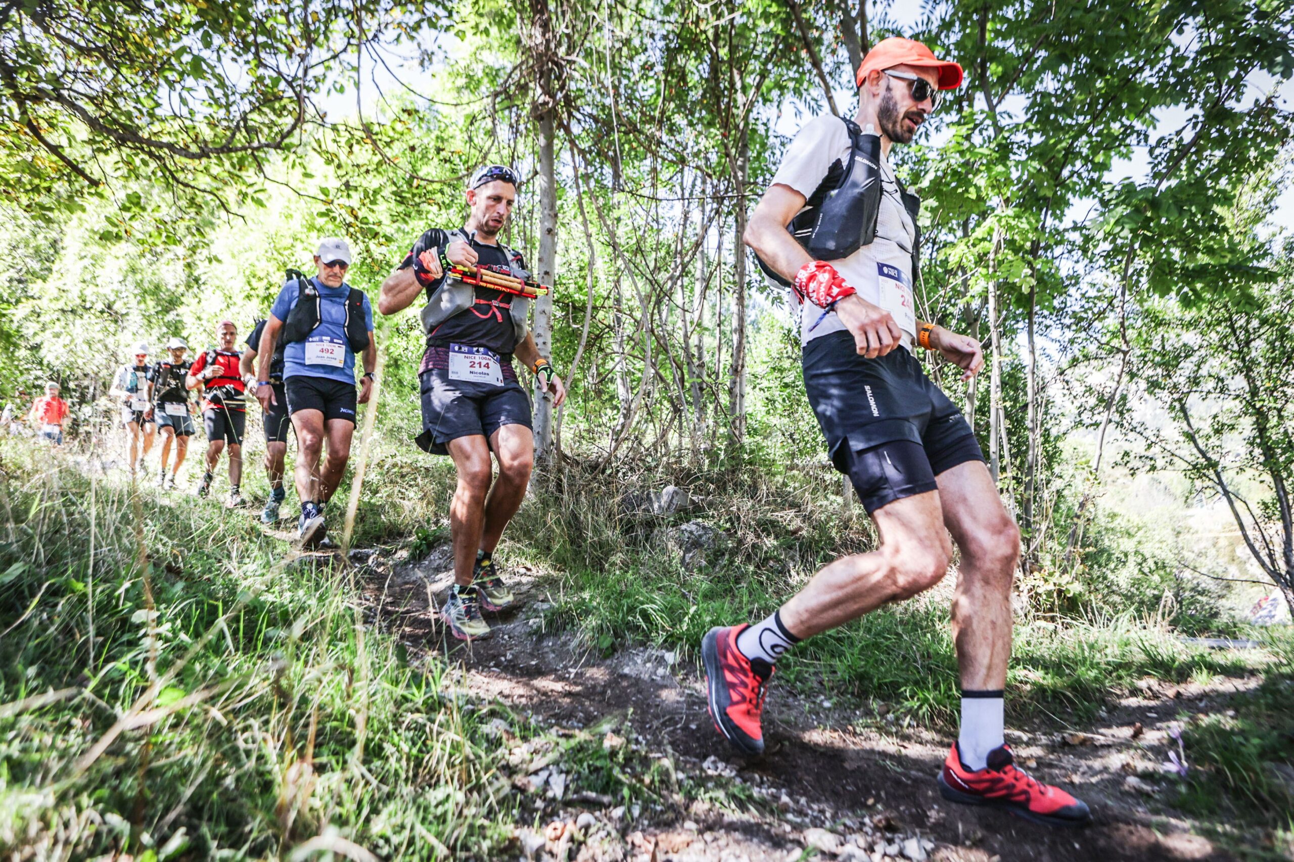 A line of men run down a trail through a forest, at the front is Pete wearing red shoes, black shorts and a white top.