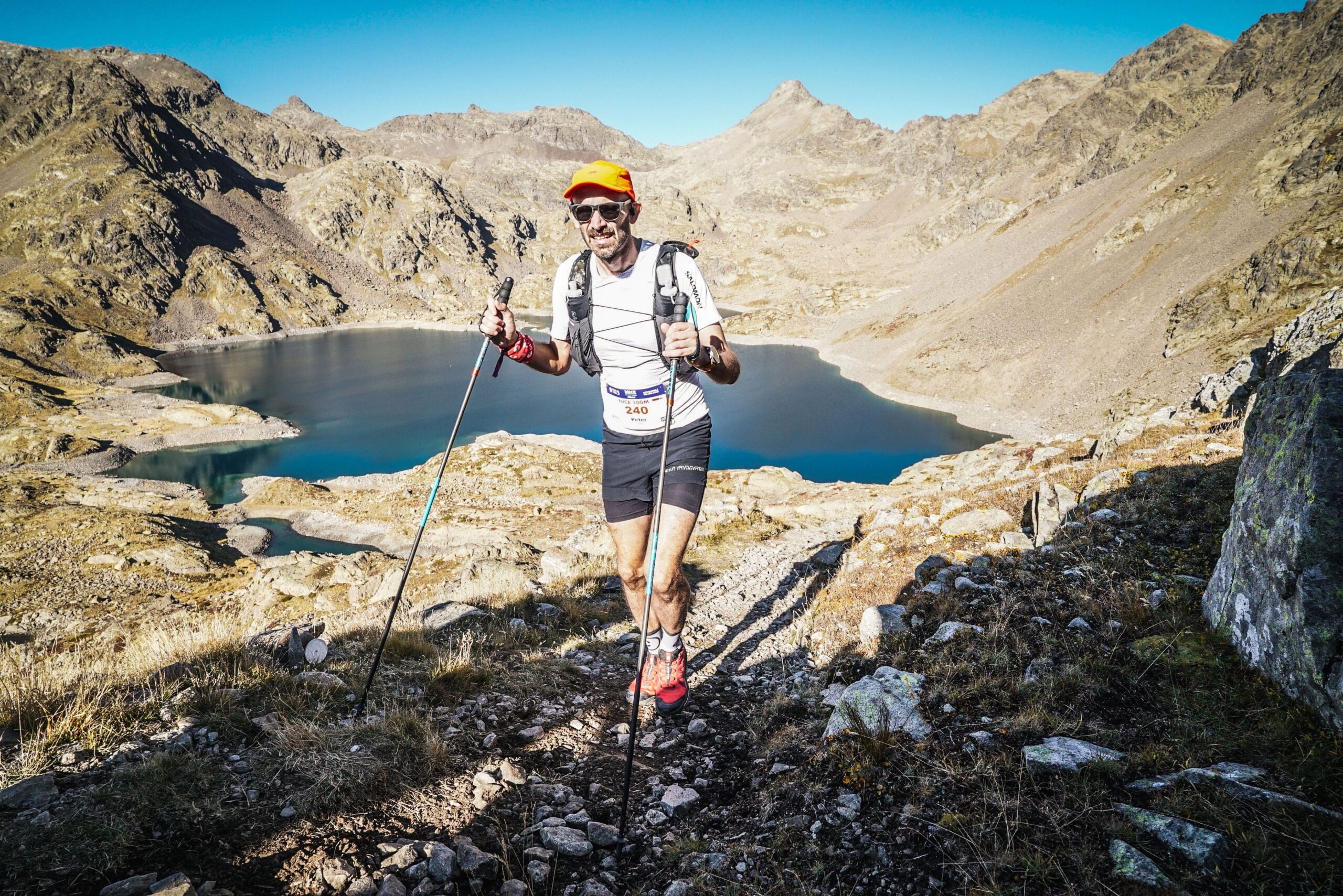 Pete runs along a rocky trail with a lake behind, framed in an ampitheatre of mountain peaks.