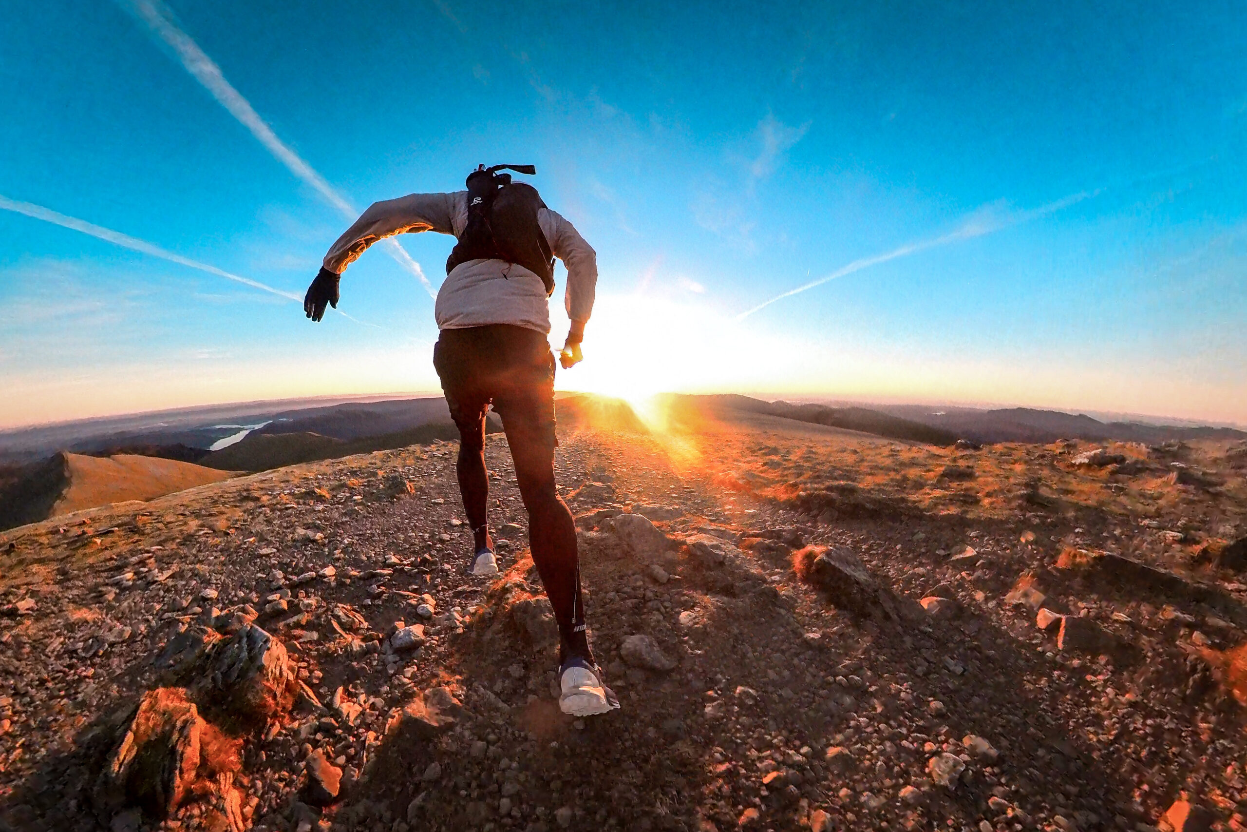 Pete running away from the camera into the sun rise on top of a mountain. He's wearing leggings and a trail pack and is at full stride, while the ground beneath him is covered in loose rocks.