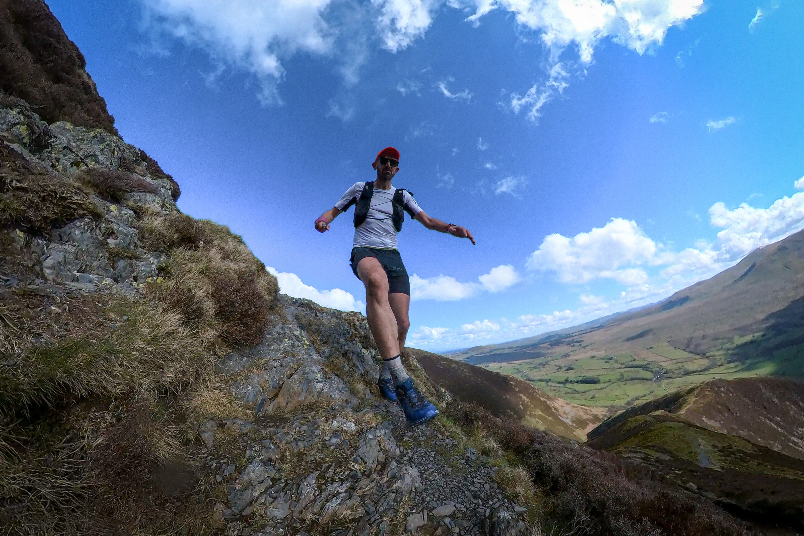 Pete is in the centre of the picture, running down hill, arms out to either side for balance, taking a big stride. The path is rocky and there is a crop of rocks to one side while the hill rolls away grassy to the other.