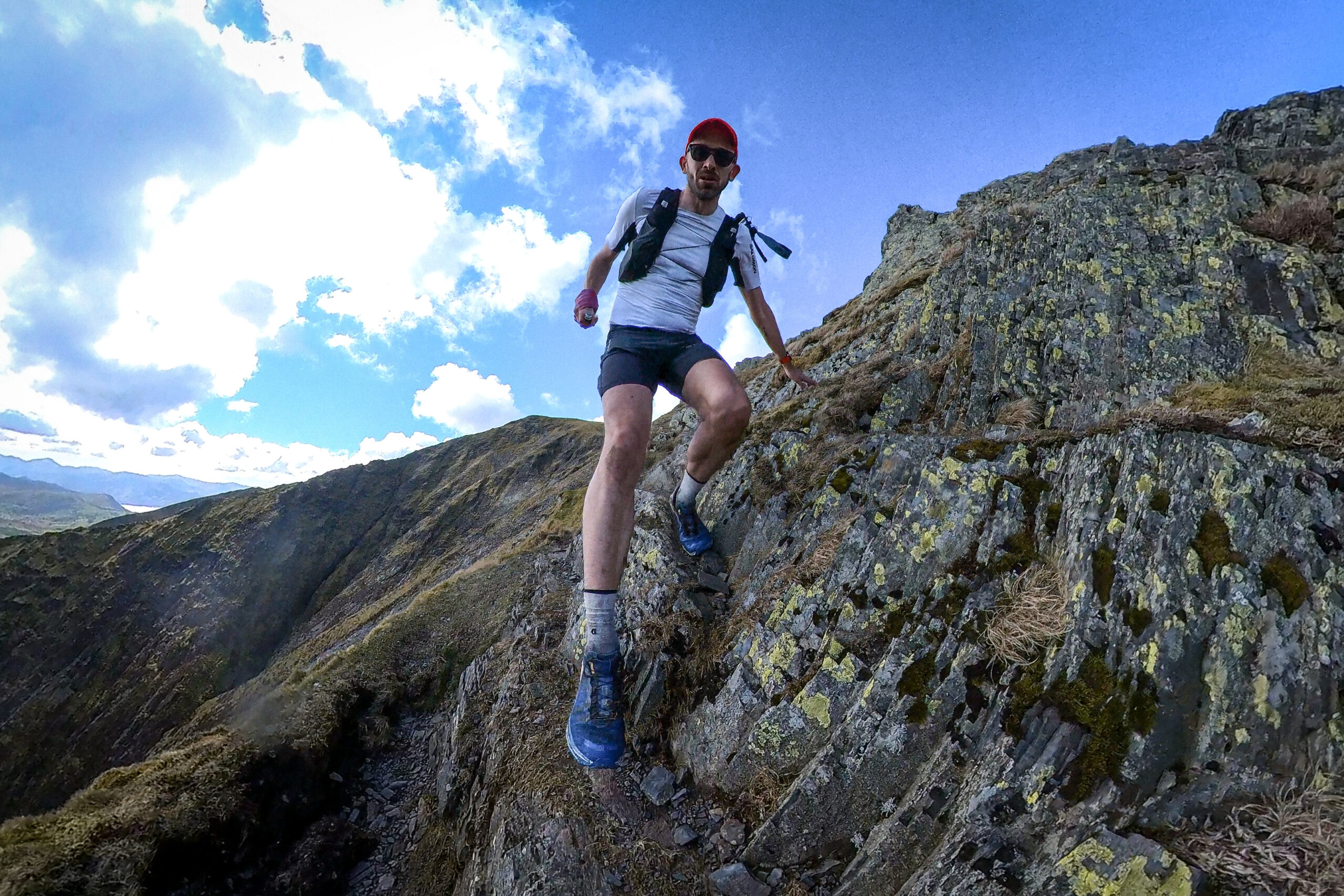 Pete runs along a mountain ridge, one hand steadying him against the rock while he steps forward on a ledge.