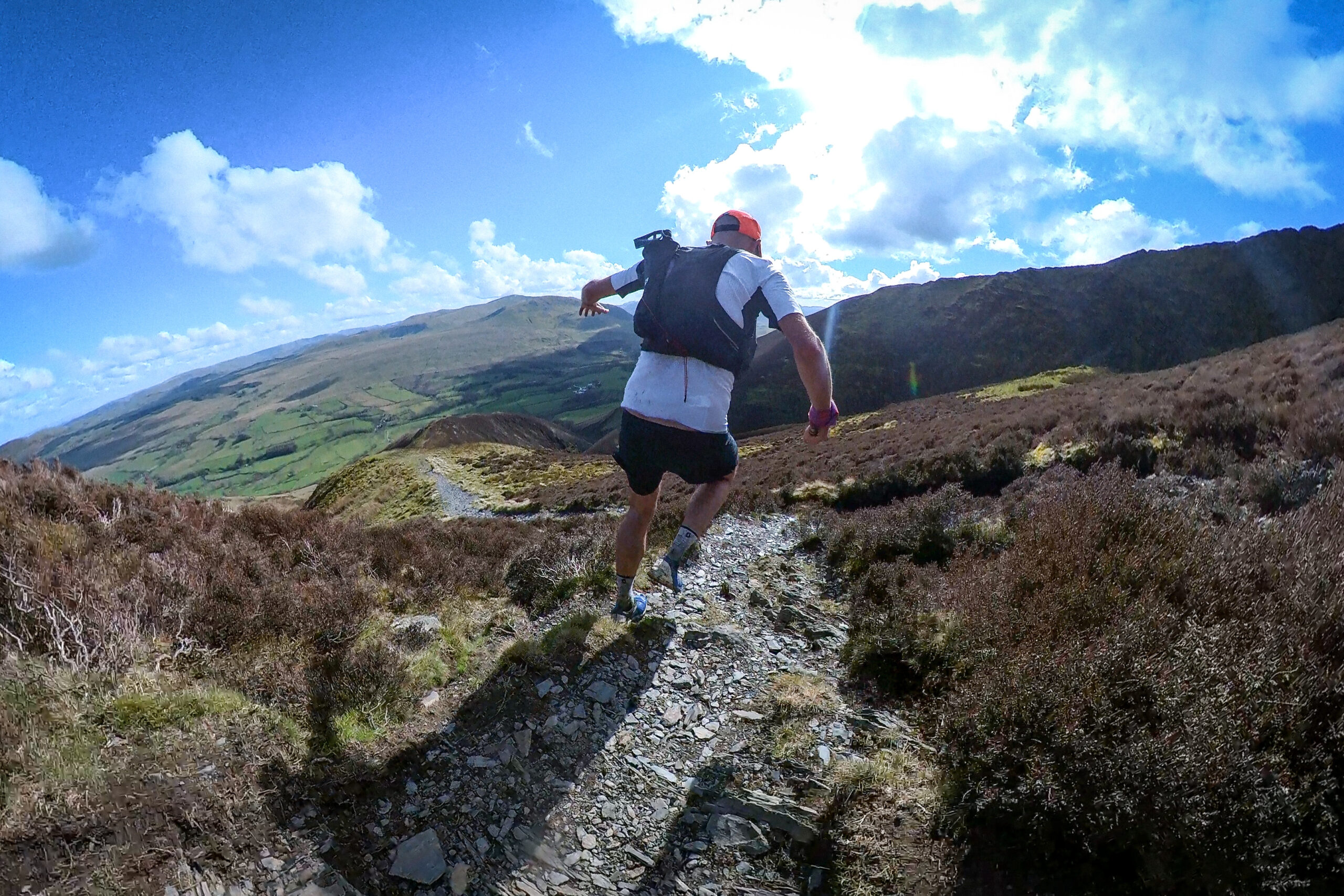 The view from behind as Pete runs down a path, on a ridge, with heather to either side of the rocky path and clear blue skies.