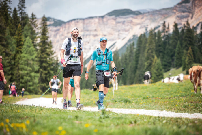 Two runners walk down a dirt track with trees and mountains behind them