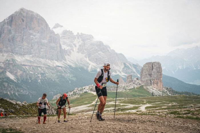A runner using trekking poles runs up hill in front of large mountains