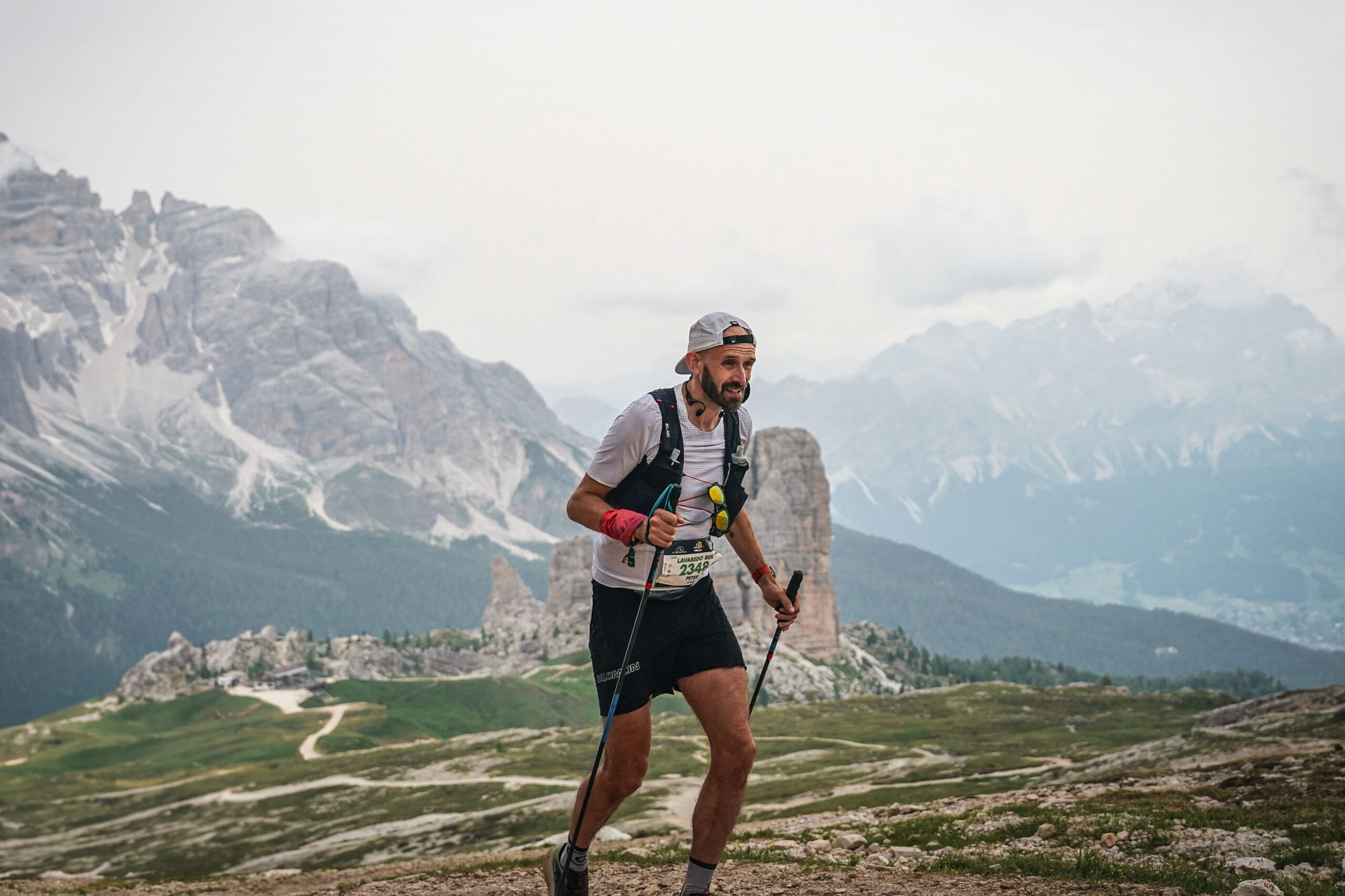 A runner uses trekking poles, moving across the front of the picture with a mountain range in the background.