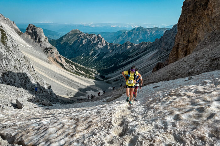 A runner wearing yellow approaches the top of a steep climb on loose stones at the end of a long valley, flanked by mountains.