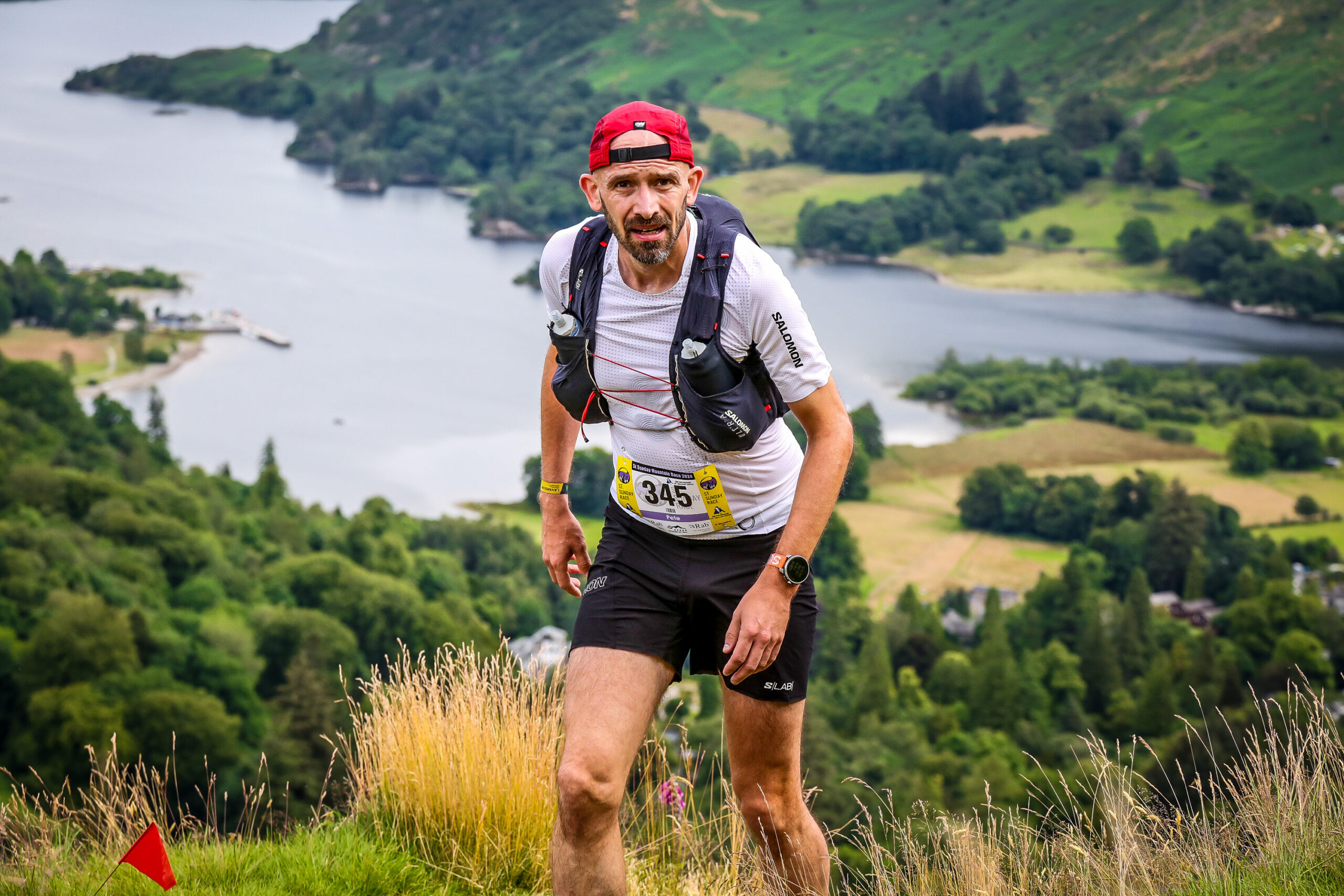 A runner with a red cap looks at the camera while running up a hill, a lake in is in the background, surrounded by tree covered slopes.