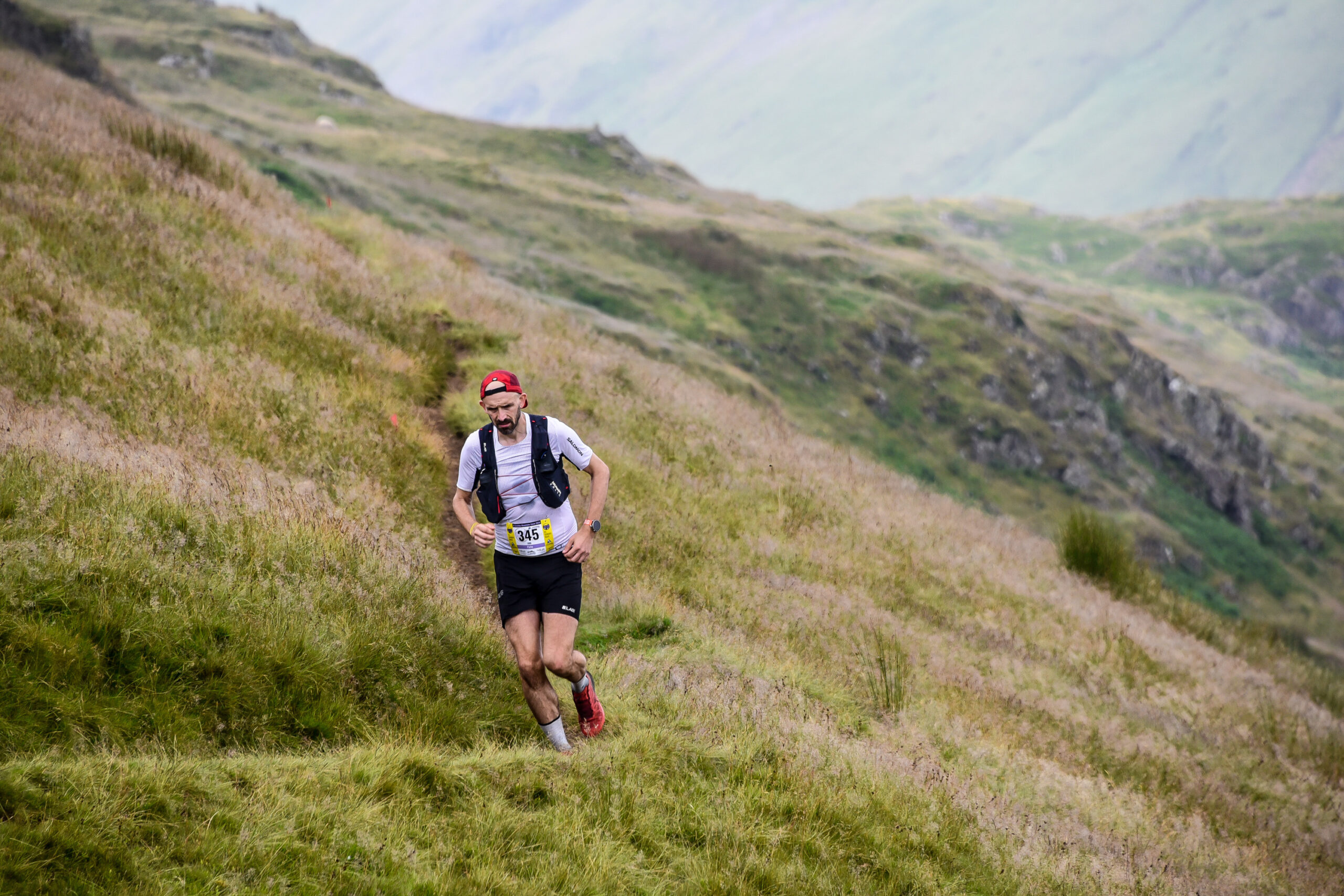A runner runs along a grassy trod on the side of a lake district fell, with mountains in the background.