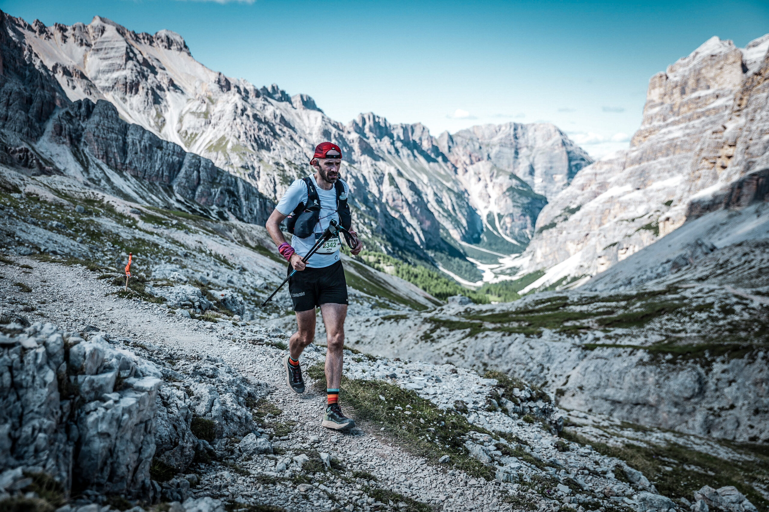 A runner runs along a mountain trail with a valley behind him flanked by high mountains.