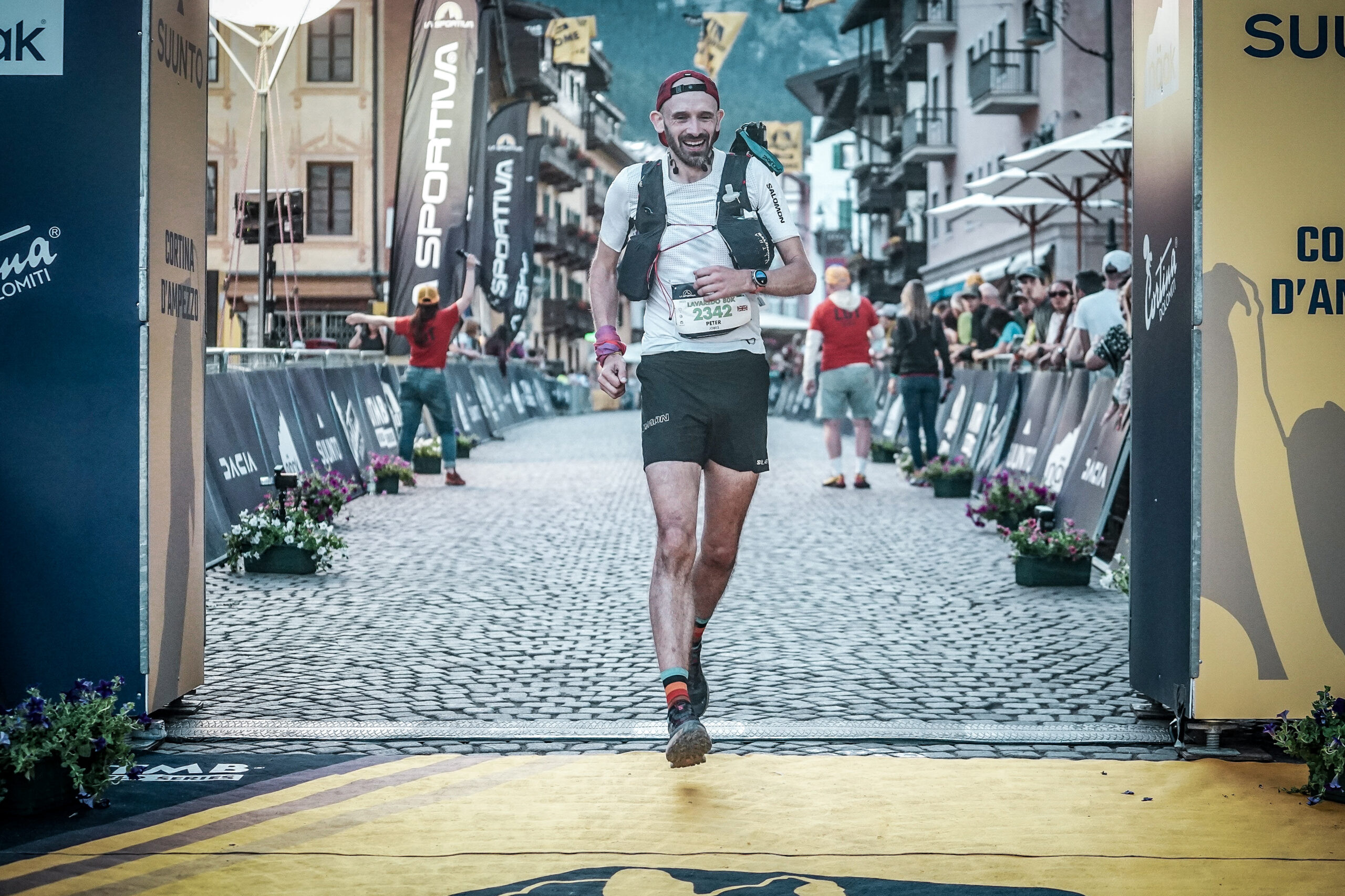 A man runs over the finish line of Lavaredo Ultra Trail, he is taking a long stride and smiling. Behind him the street is full of flags and hoardings for the event.