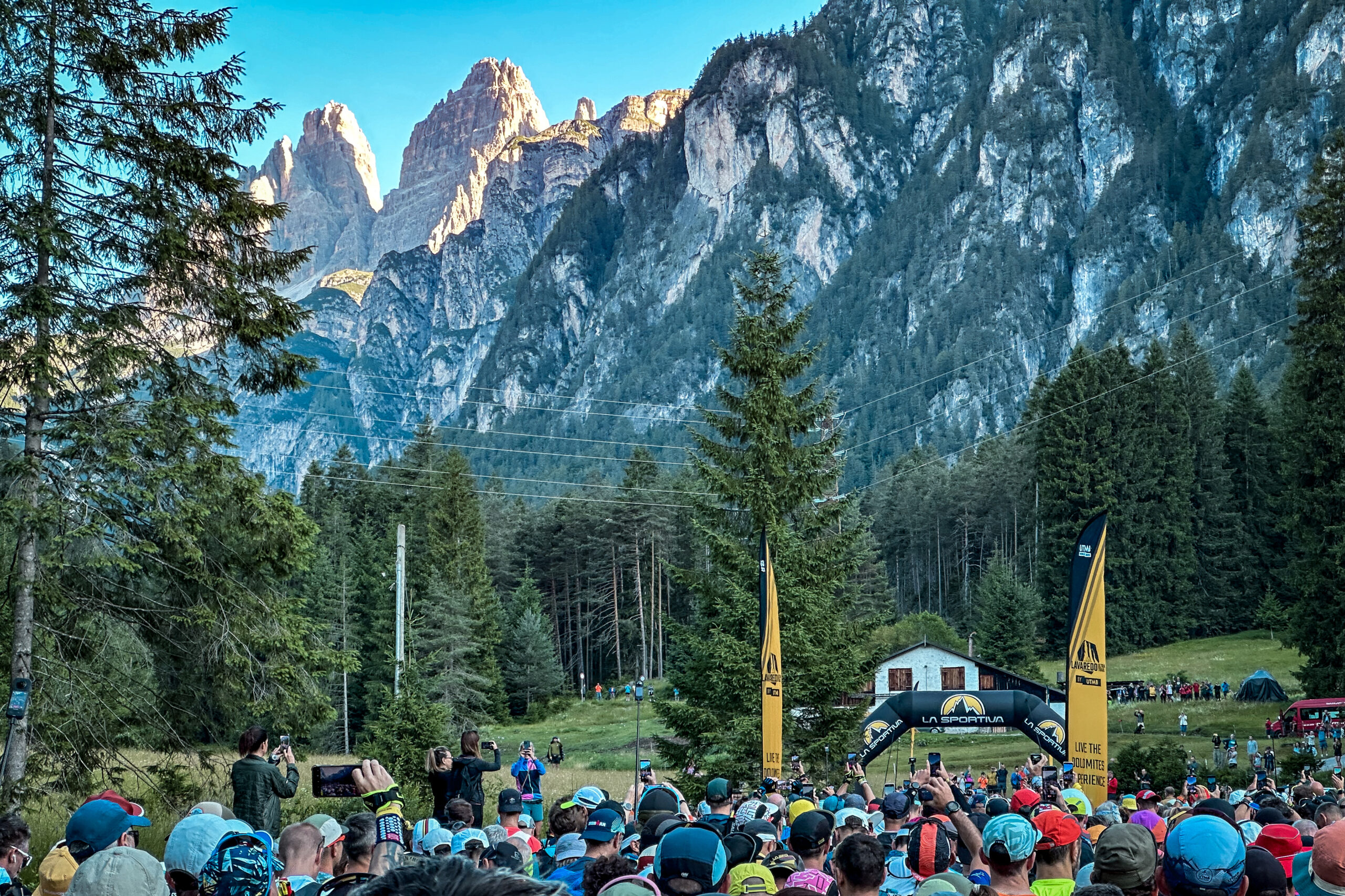 People crowd around the start line to a race, with a backdrop of forest and mountains and three distinct peaks, Tres Cime, poking out behind the other mountains