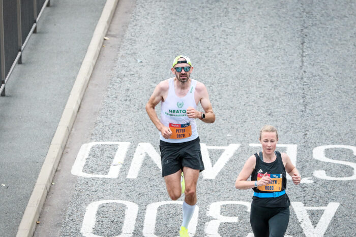 Two runners run along the road, a lady in black in front with a man behind in a white vest and sunglasses.