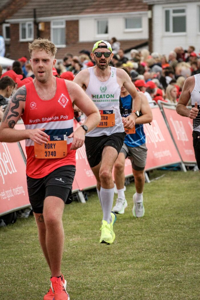 Two men run towards the finish line of the Great North Run. Rory, in front of me, runs in a red North Shields Poly vest. I am behind in a white Heaton Harriers vest, mouth open and both feet off the ground.