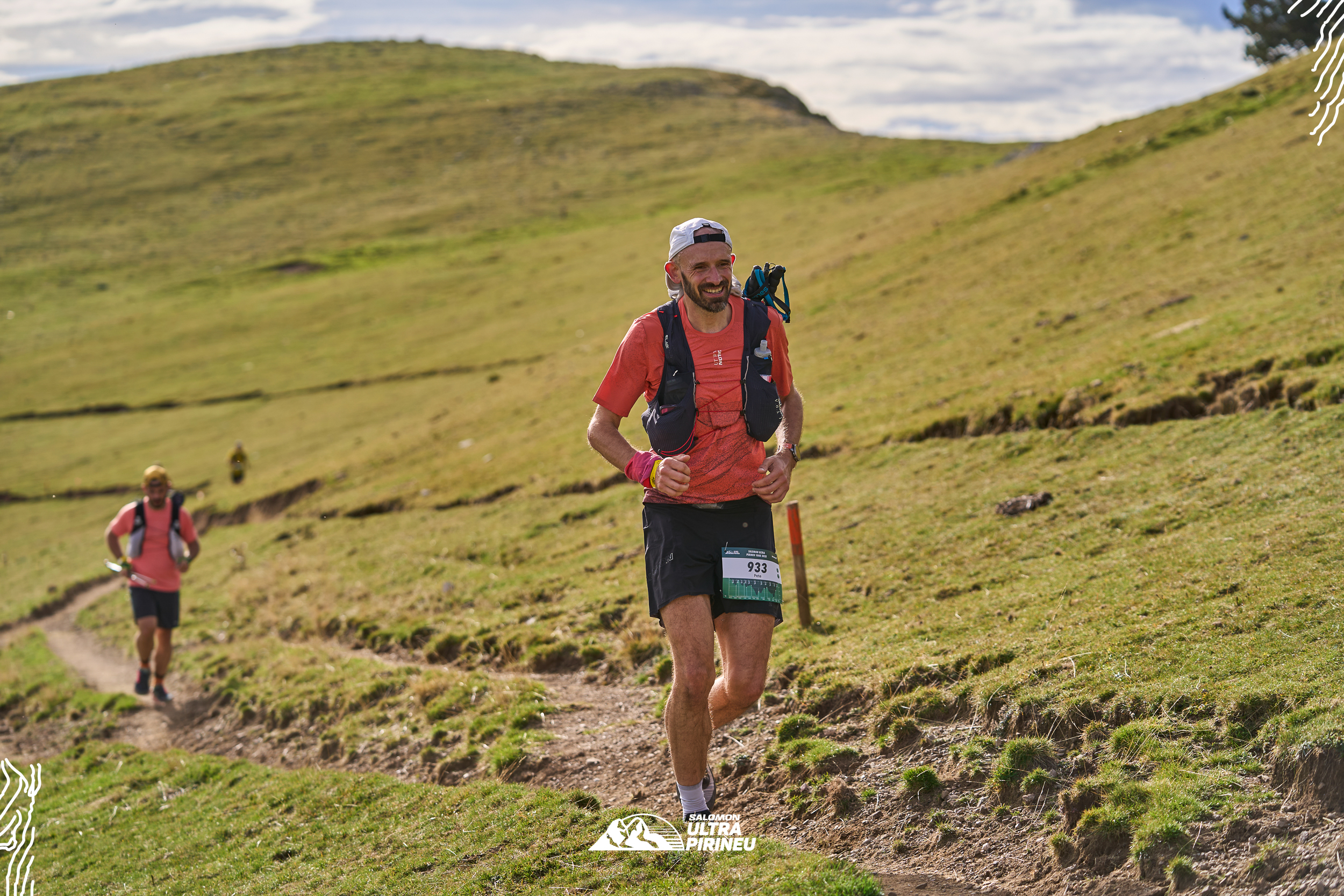 A runner in a red t-shirt runs along a trail on a grassy hillside