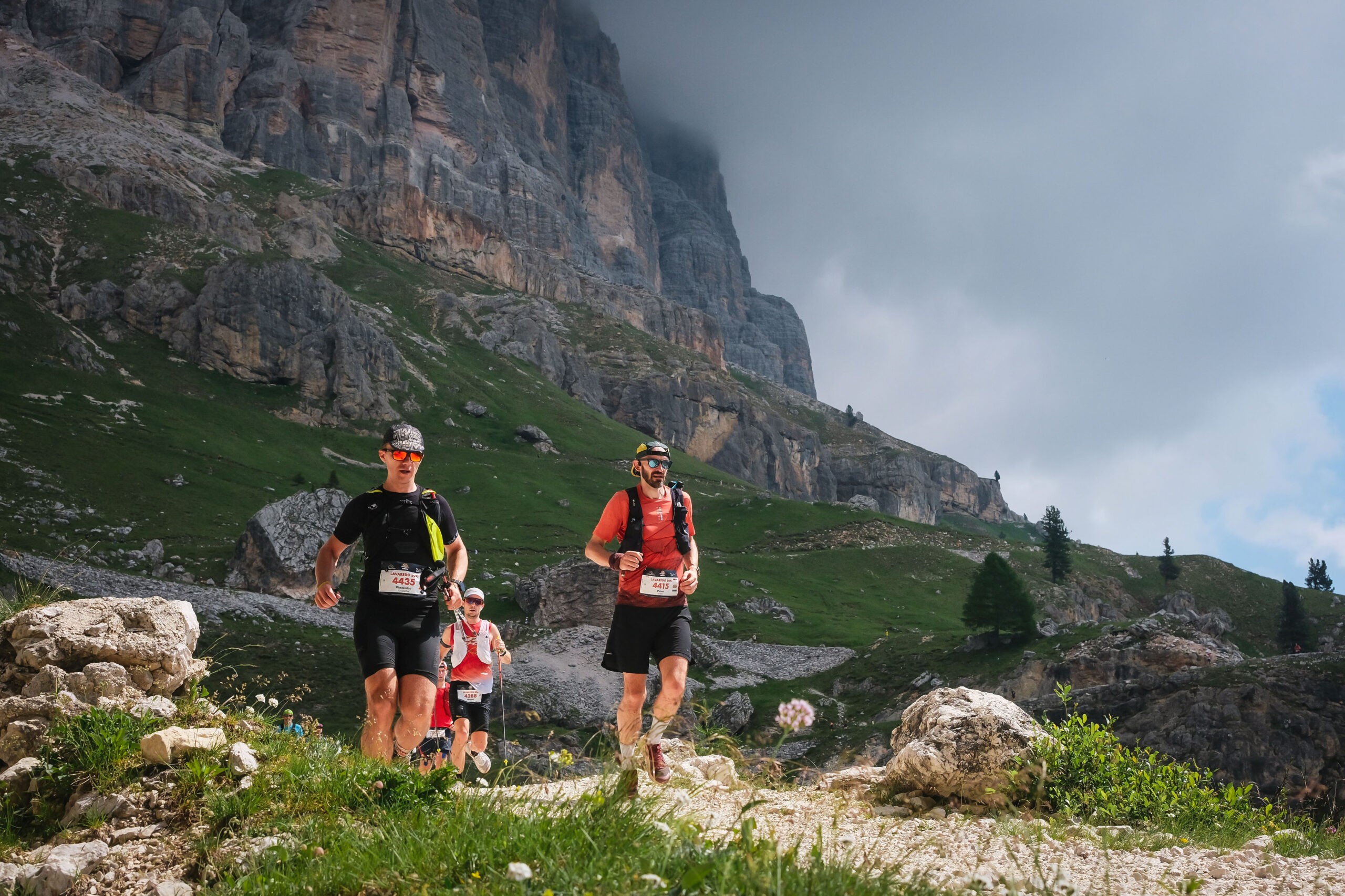 Three runners run down a dirt trail in front of a mountain, clouds cloak the mountain. One runner has just overtook another.