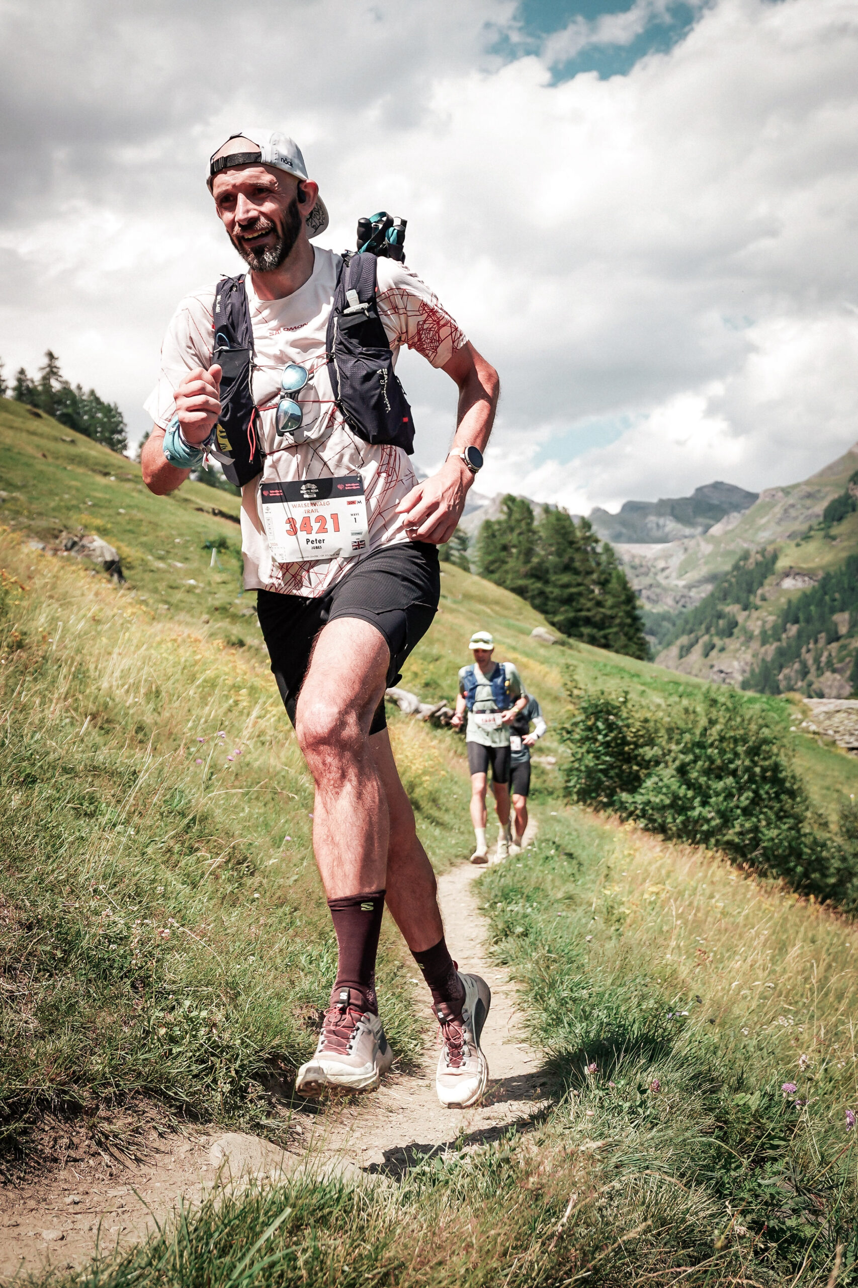 A runner runs along a single track trail, two more runners are visible in the background