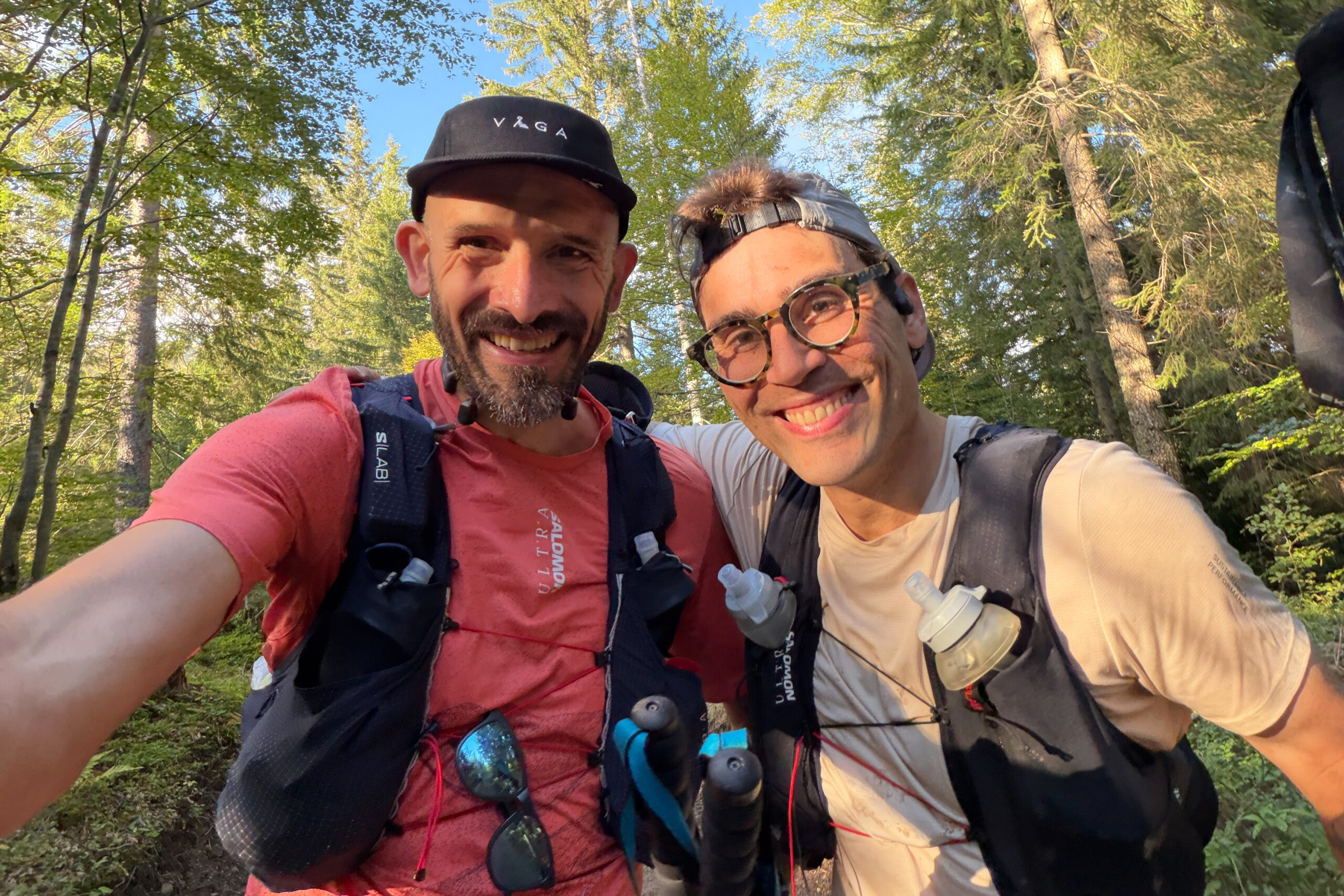 Two men smile at the camera in a selfie taken mid-trail race, they're wearing running clothes and hydration vests and stood in a forest.
