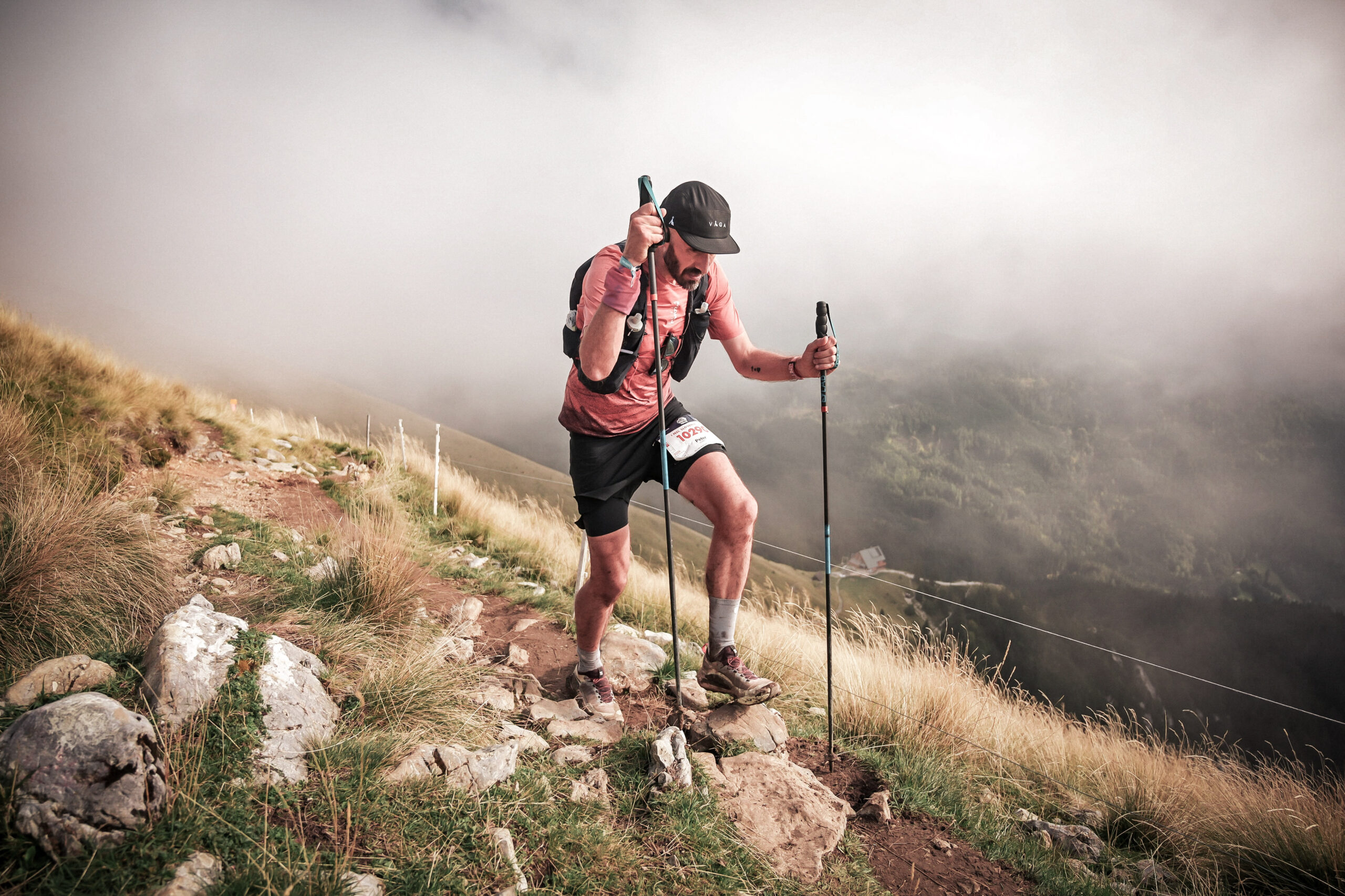 A trail runner struggles up a climb using trekking poles, with misty mountains in the background.